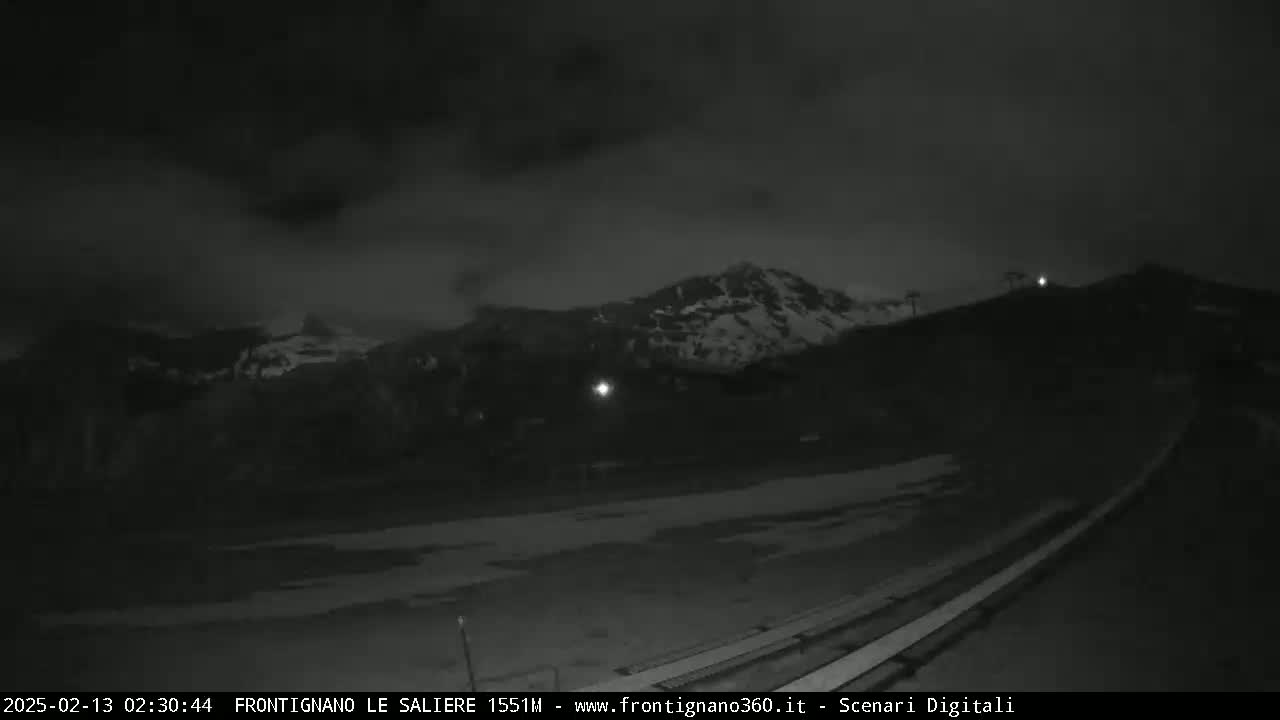 A nighttime, overcast view shows snow-capped mountains in the distance, a flat, snow-covered area in the foreground, and a long, thin structure leading toward the mountains.