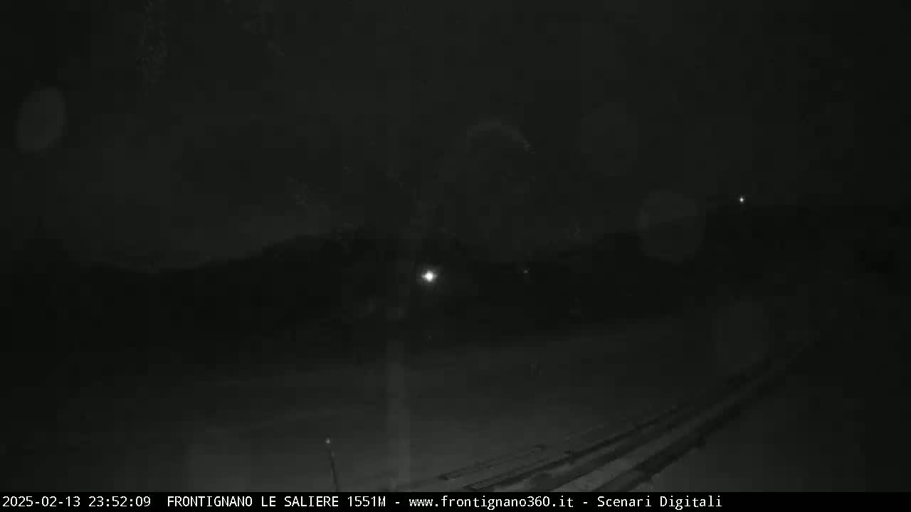 A nighttime, snowy scene shows a dark landscape with faint lights in the distance.