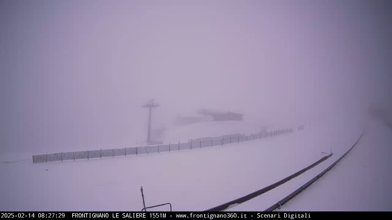 A snow-covered landscape is obscured by heavy fog, with a ski lift and building barely visible in the distance.