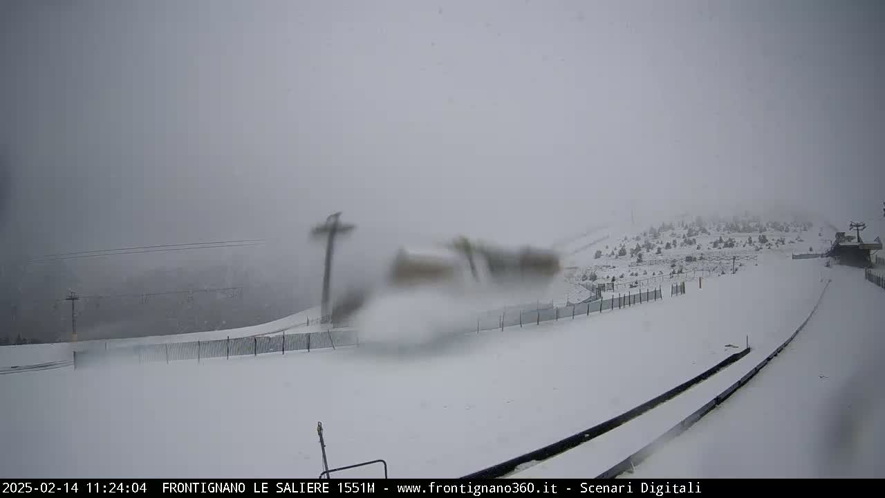 A snow-covered ski slope is visible through a snowy, foggy atmosphere, with ski lifts and sparse trees in the background.