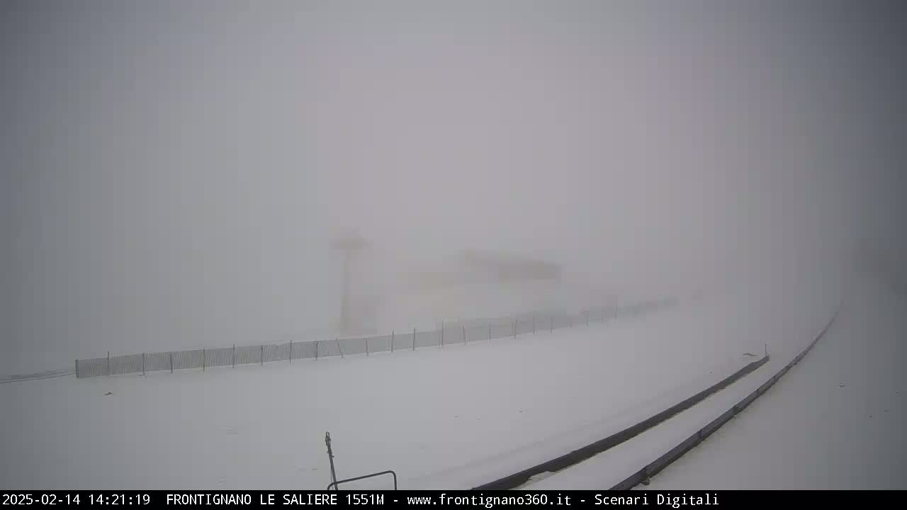 A snow-covered landscape with a fence and indistinct structures in the distance is shrouded in heavy fog or blowing snow.
