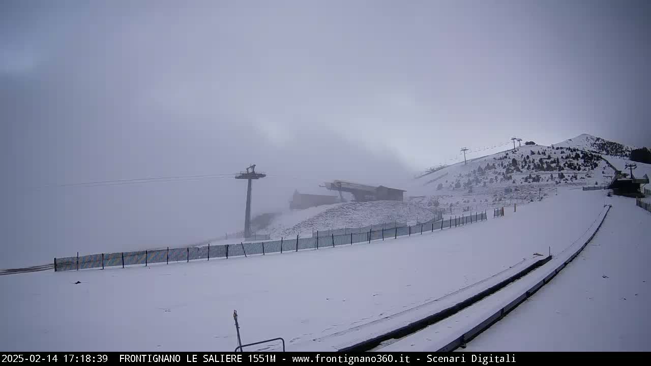 A snow-covered ski slope with chairlifts and a building is visible under foggy conditions.