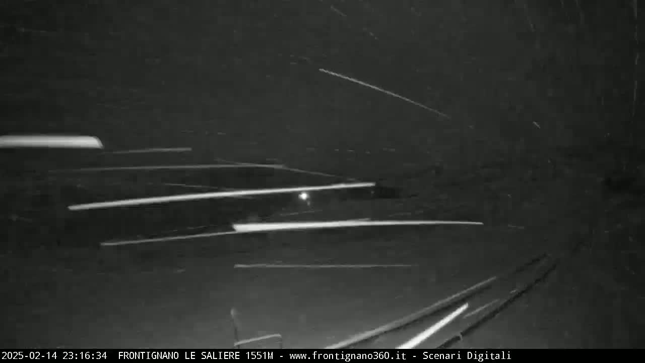 Streaks of light from vehicles are visible on a dark roadway at night during a snowfall.