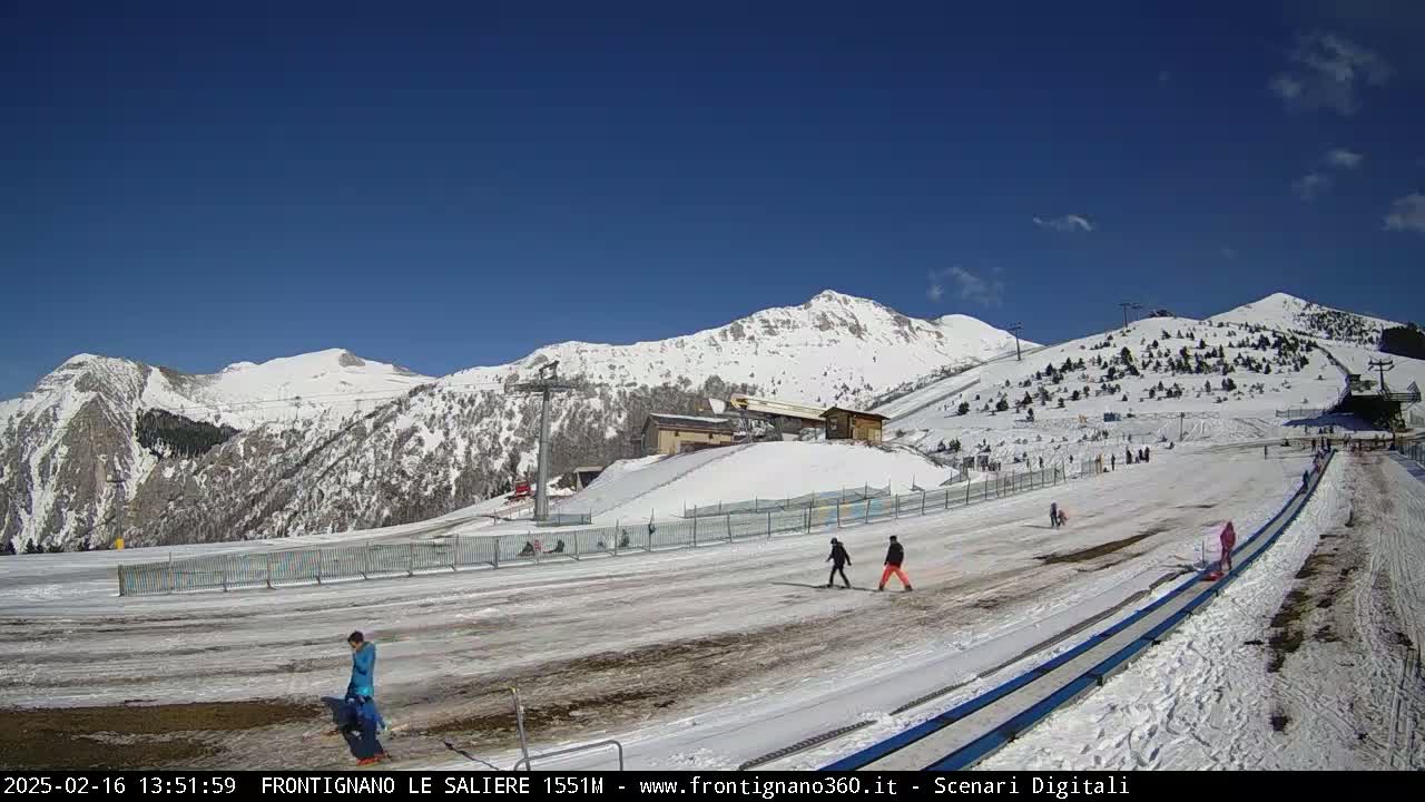A snow-covered mountain slope with a few people walking and a building in the background under a clear blue sky.