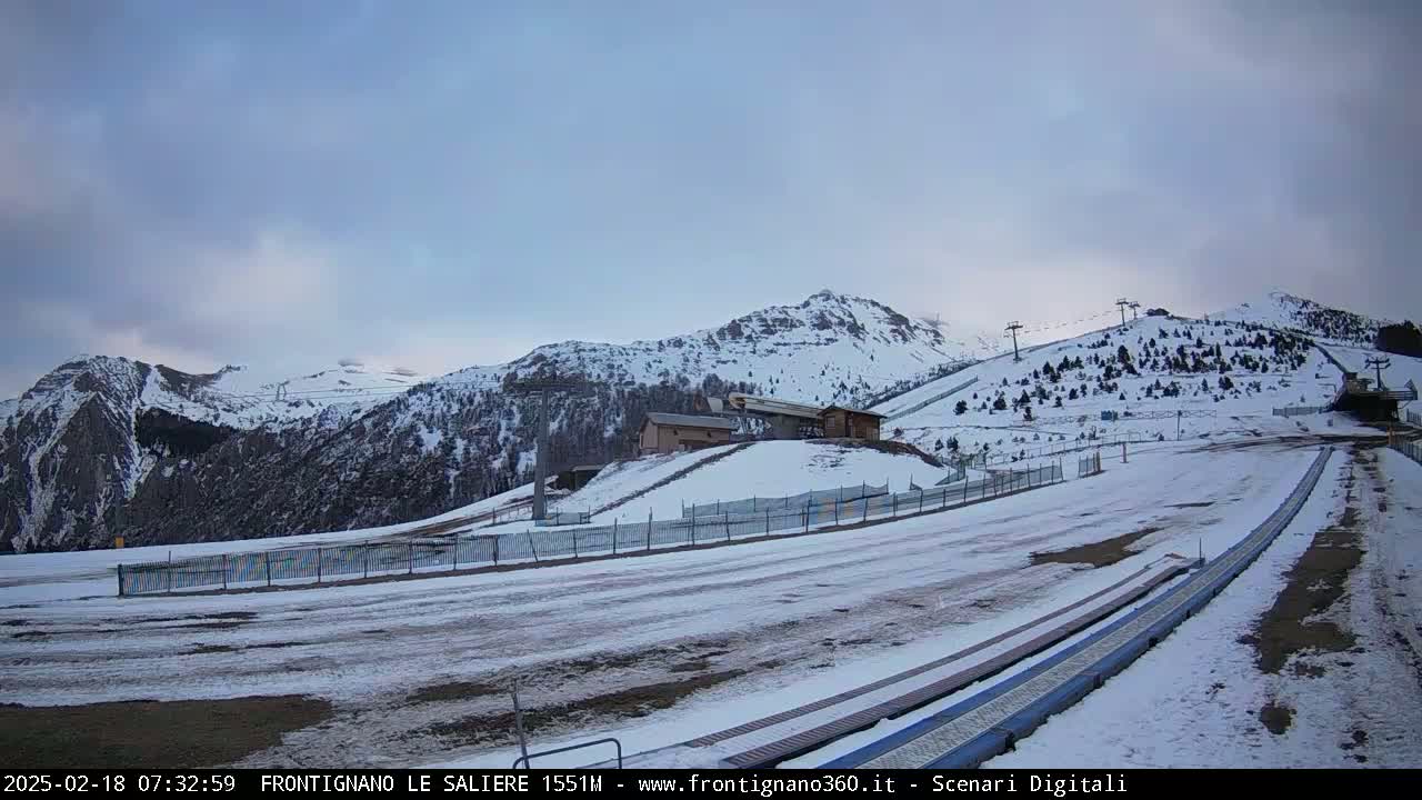 A snow-covered mountain landscape with ski lifts and a building, under a cloudy sky.