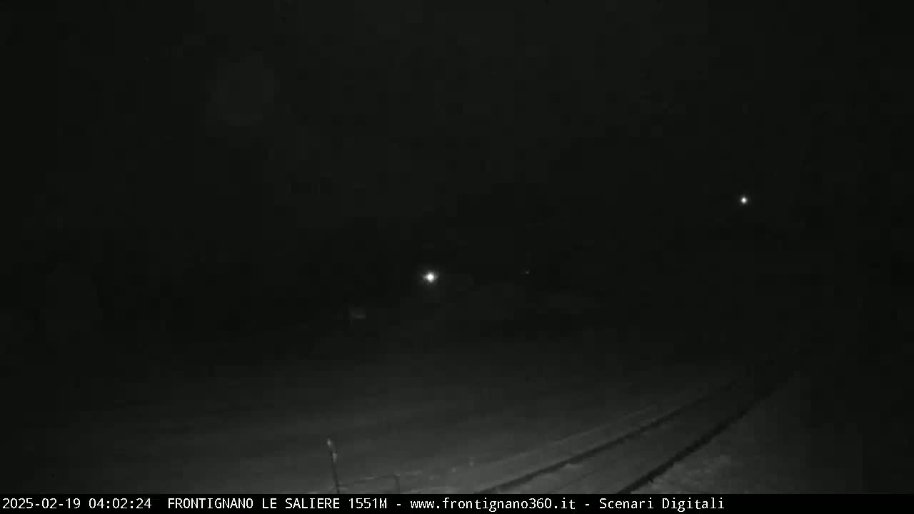 A dark, nighttime outdoor scene shows a road with tire tracks, faintly illuminated by distant lights under a mostly clear sky.