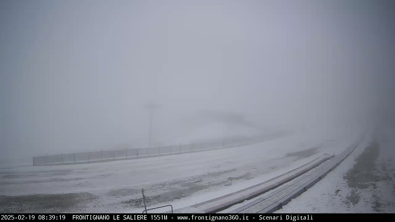 A snow-covered landscape with a fence and tracks is barely visible through a thick fog.