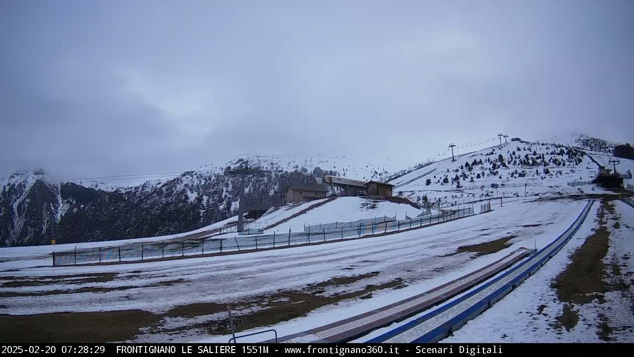 A snow-covered ski slope with a lift and buildings in the background under an overcast sky.