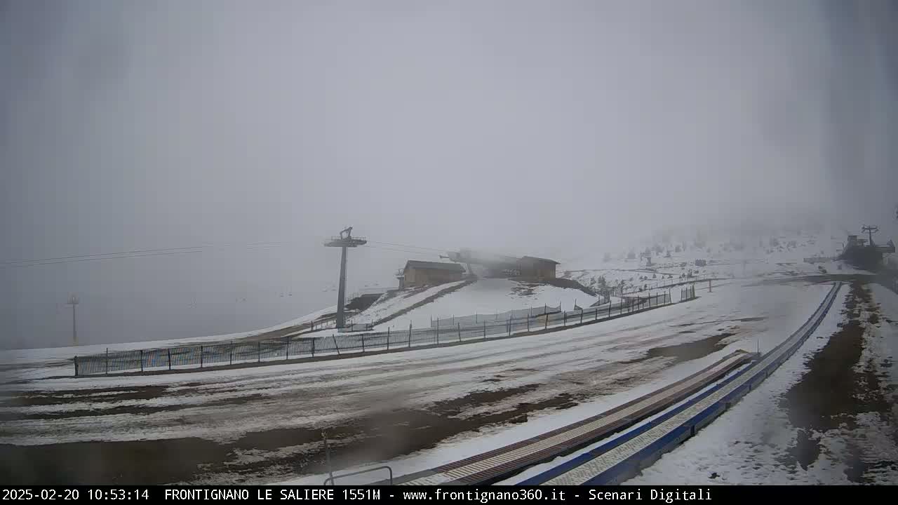 A foggy, snowy scene shows a ski lift, small buildings, and a snow-covered path leading to what appears to be a ski run.