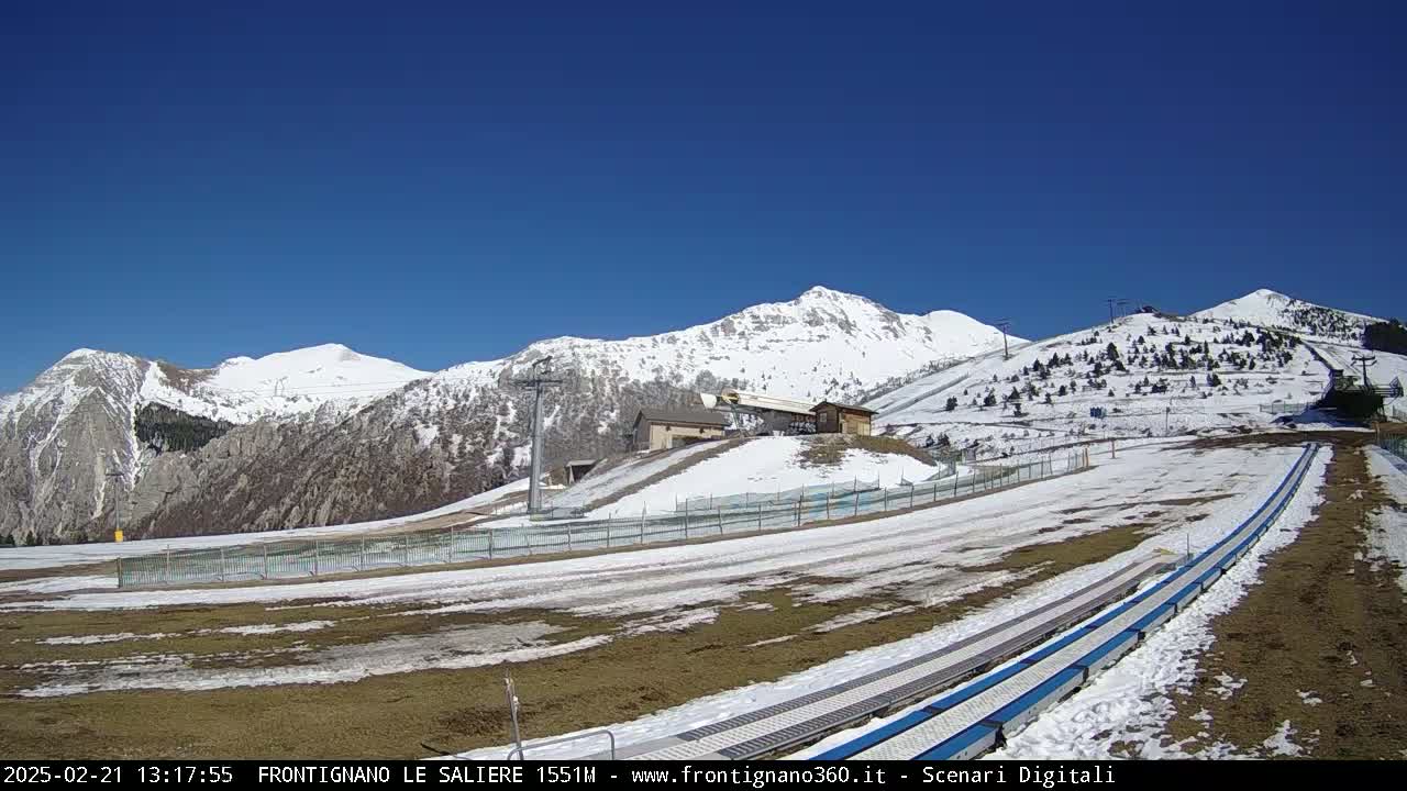 A snow-covered mountain range is visible under a clear blue sky, with patches of snow and bare ground at the base of the mountains, featuring a ski lift and what appears to be a snow tubing run.