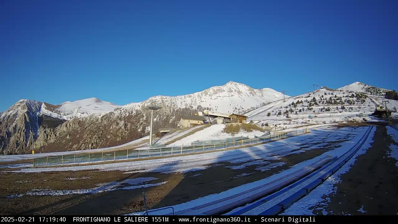 A snow-dusted mountainside ski area with a clear blue sky, featuring a ski lift and a run with some patches of snow.
