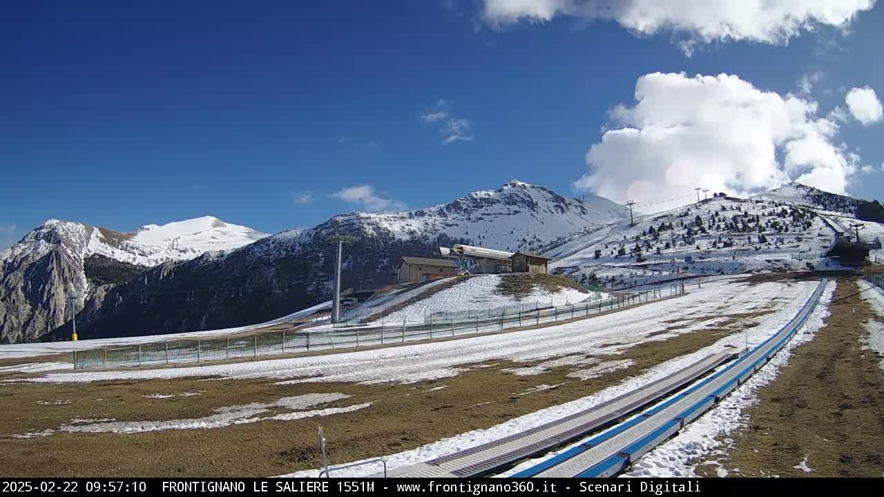 A mostly sunny day reveals snow-capped mountains, ski lifts, and a snow-covered ski run with a snow conveyor belt in the foreground.