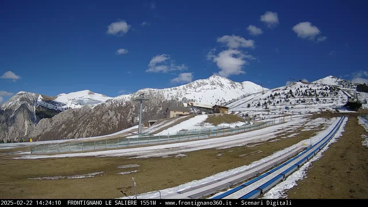 A mostly sunny day reveals snow-capped mountains and a ski slope with snow patches and some grassy areas, featuring a chairlift and a small building.