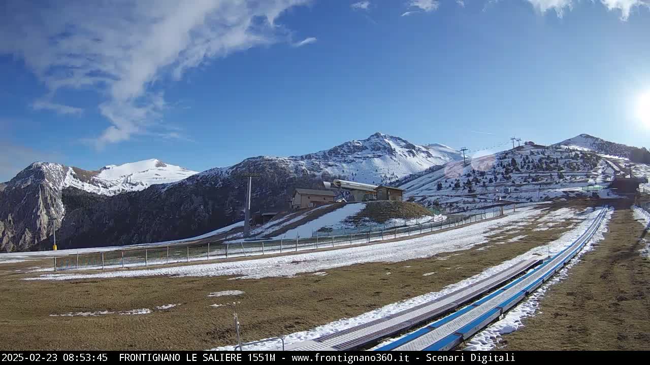 A mostly sunny day reveals snow-capped mountains, ski lifts, and a snow-covered, grassy area with two long, parallel tracks leading towards a building at the base of the mountains.