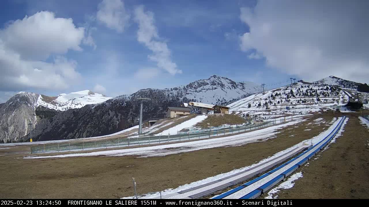 A partly cloudy day reveals snow-dusted mountains and ski lift infrastructure, with snow patches on the ground and what appears to be a ski run or snow tubing track in the foreground.