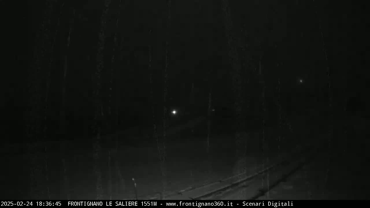 A dark, nighttime scene shows a dimly lit roadway with what appears to be rain or snow falling.
