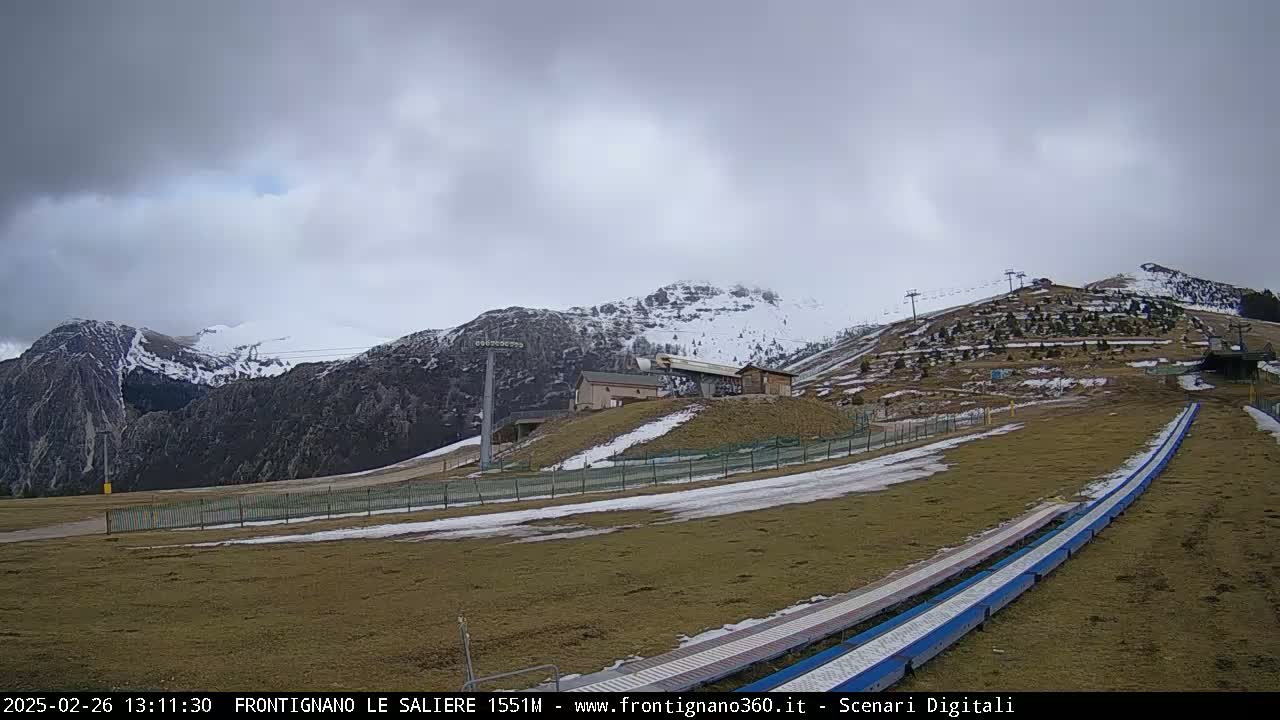A mostly cloudy day reveals snow-dusted mountains and hills, with a ski lift and grassy areas in the foreground.