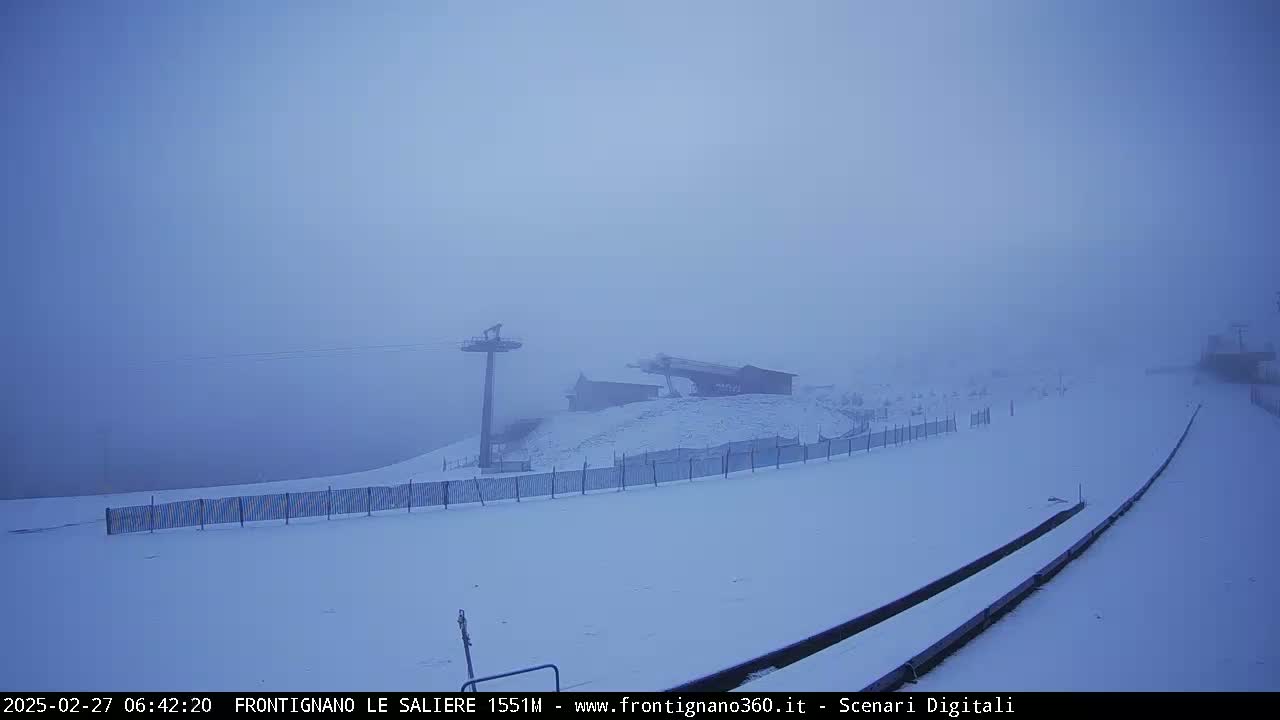 A snow-covered ski resort is shrouded in thick fog, with a chairlift and buildings barely visible in the distance.