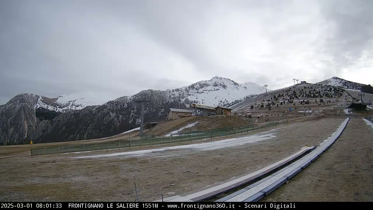 A mostly cloudy day reveals snow-dusted mountains, ski lifts, and a ski run with snow-covered patches in a mountainous area.
