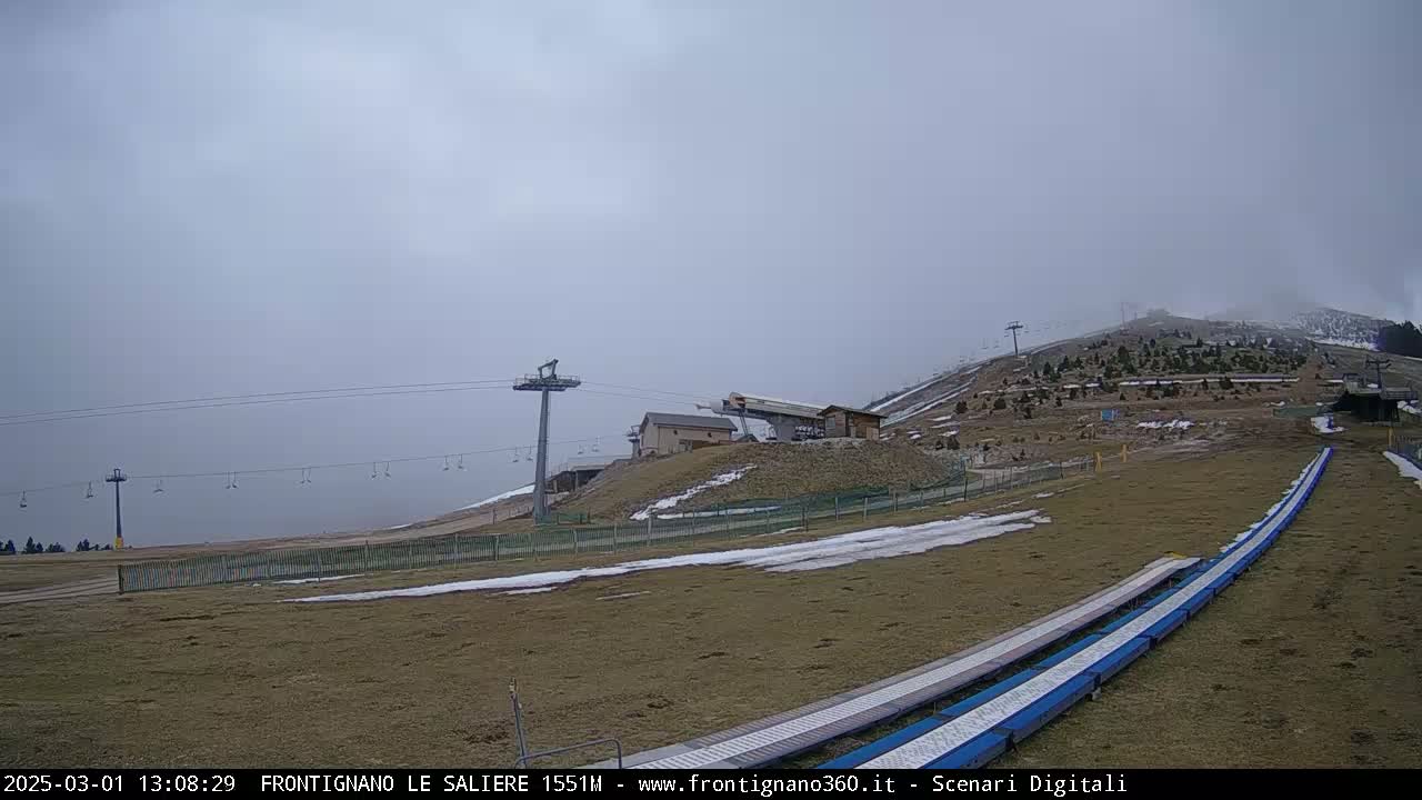 A foggy, overcast day at a ski resort shows a chairlift, a ski lift station, and two long snow slides on a sparsely snow-covered hillside.