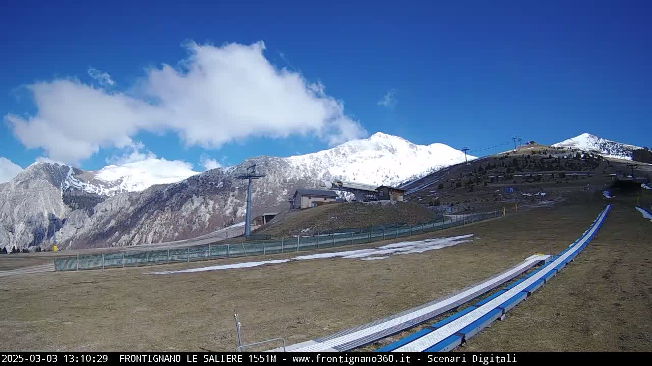 A mostly sunny day reveals snow-capped mountains in the background, with a ski lift and grassy area in the foreground containing two snow tubing tracks.
