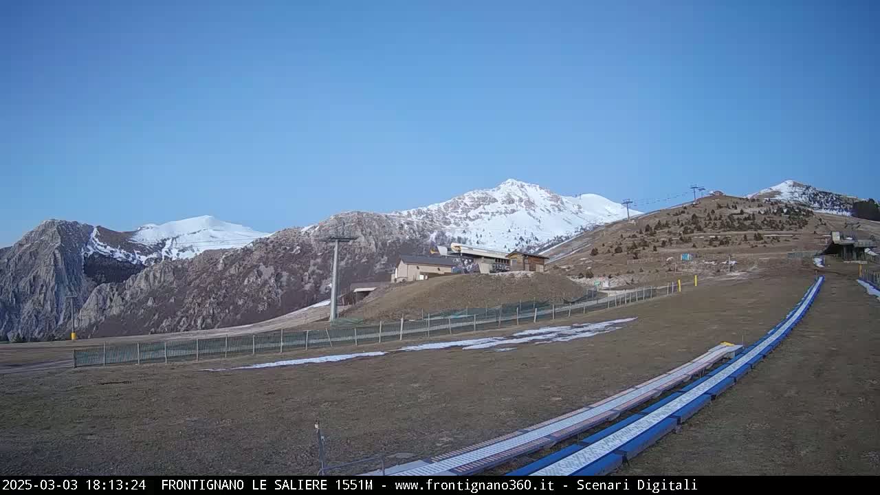 A snow-capped mountain range is visible under a clear blue sky, with a ski lift and slope in the foreground.