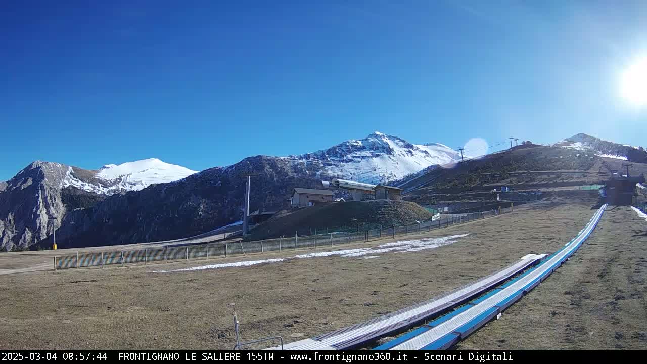 A sunny, snow-capped mountain range is visible behind a ski lift and snow tubing runs on a mostly brown, grassy hill.