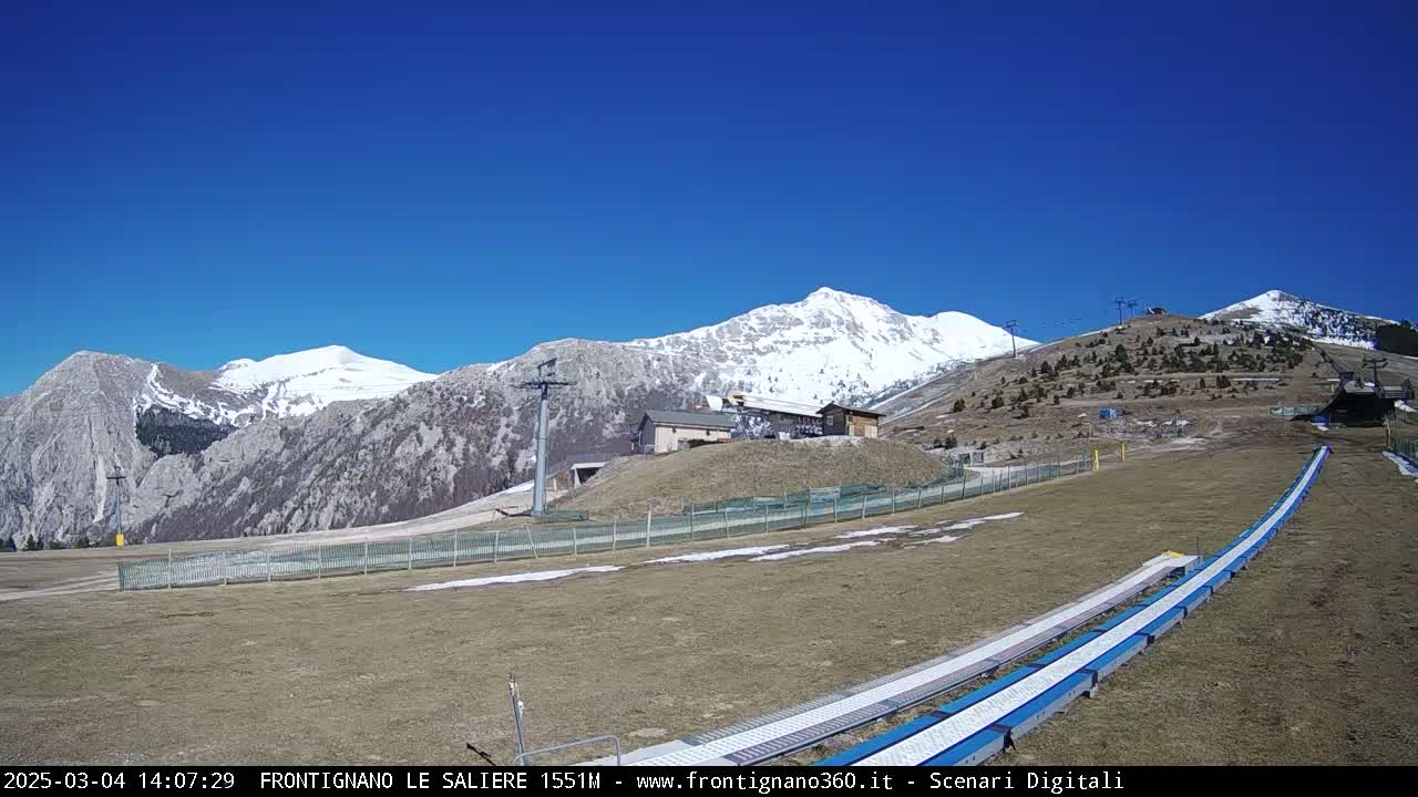 A snow-capped mountain range is visible under a clear blue sky, with a grassy area in the foreground featuring a ski lift and what appears to be a snow tubing or sledding run.