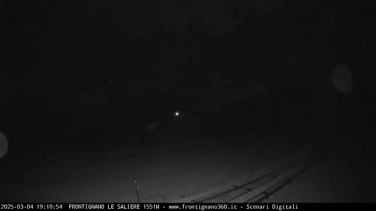 A nighttime, low-light image shows a dark, mostly featureless scene with a faint light source in the middle distance and some faint horizontal lines in the foreground.