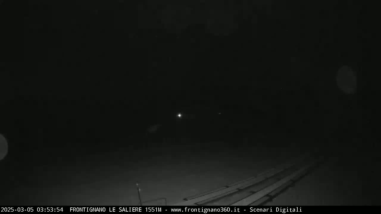 A nighttime, low-light view shows a dark, empty area with a few faint lights in the distance and what appears to be bleachers in the lower portion of the frame.