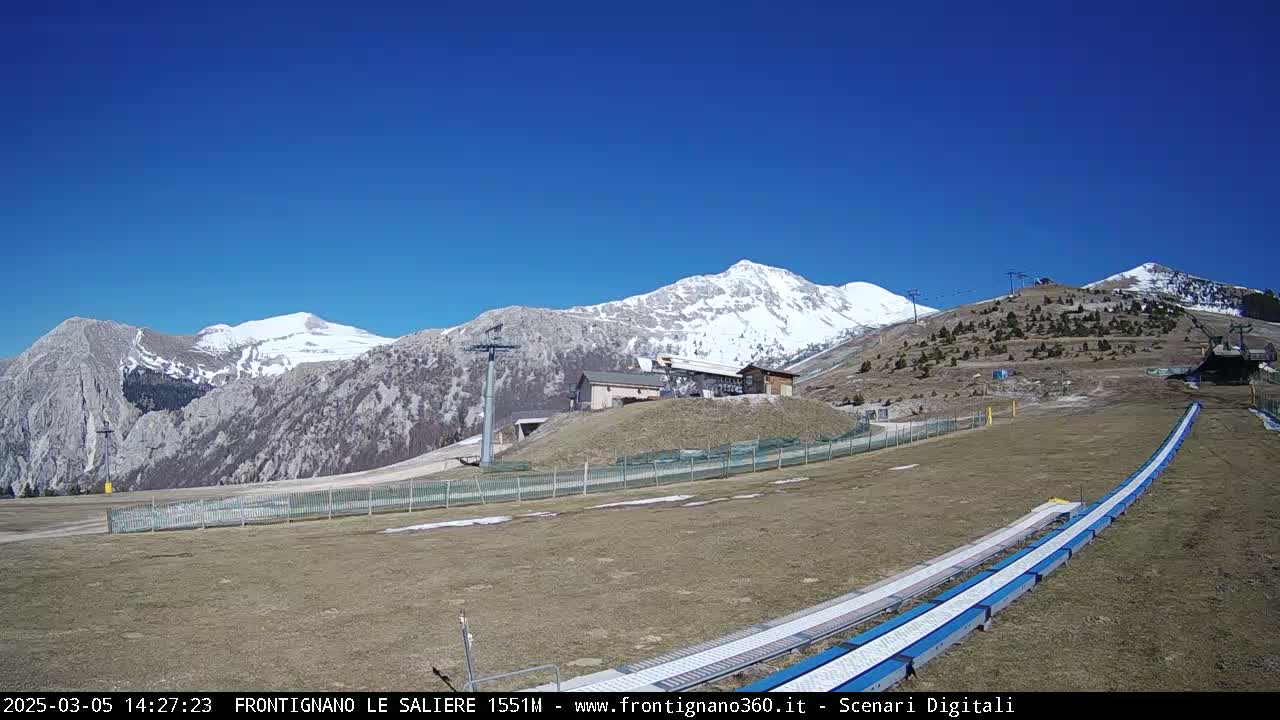 A snow-capped mountain range is visible under a clear blue sky, with a ski lift and a grassy area containing a snow tubing run in the foreground.
