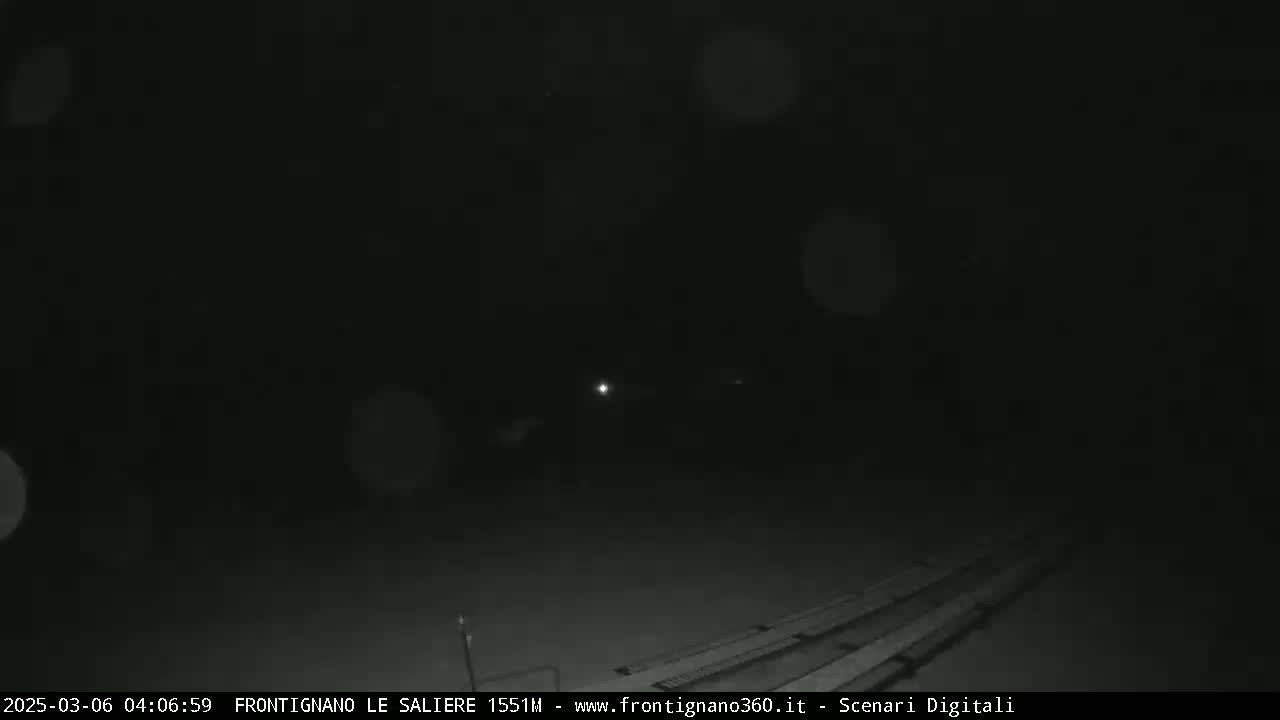 A nighttime, low-light outdoor scene shows a dark area with a faintly visible structure and a few bright lights in the distance, possibly under snowy or dusty conditions.