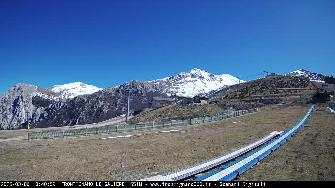 A snow-capped mountain range is visible under a clear blue sky, with a ski lift and a snow-covered conveyor belt in the foreground.