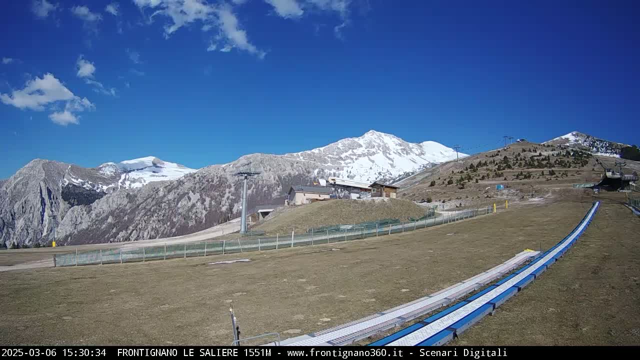 A mountain range with snow-capped peaks is visible under a mostly clear blue sky, with a ski slope and lift infrastructure in the foreground.
