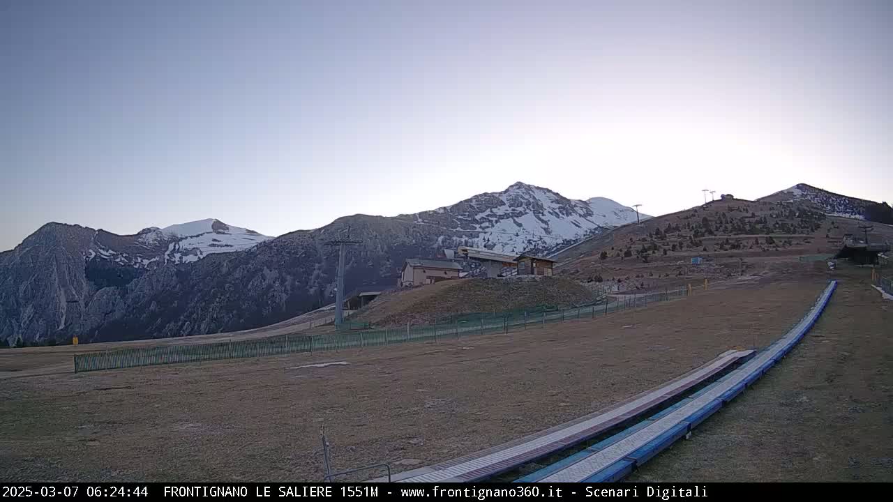 A snow-capped mountain range is visible under a clear, light-blue sky at dawn, with ski lifts and slopes in the foreground.