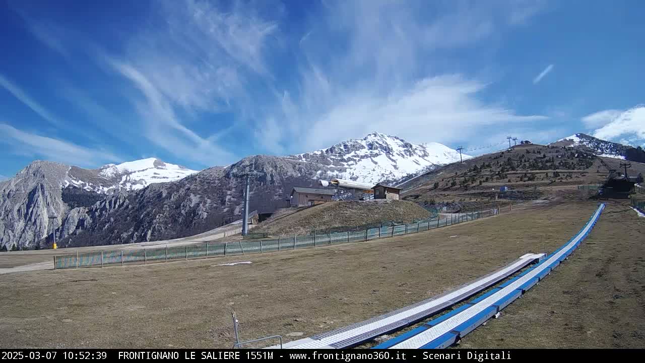 A ski lift and snow-covered mountains are visible under a mostly clear blue sky with wispy clouds,  in a dry, grassy area with a snow tubing track.