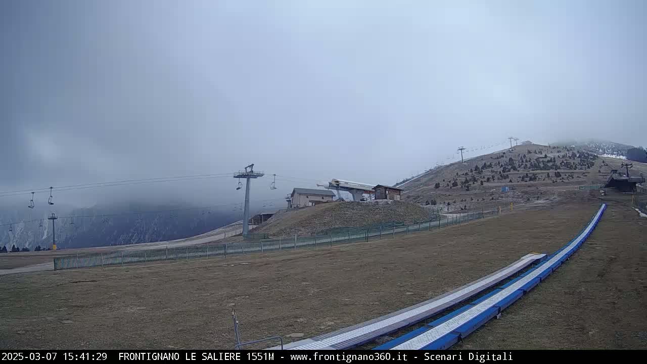 A foggy mountaintop scene shows a ski lift, a ski slope with two conveyor belts, and sparsely vegetated hills.