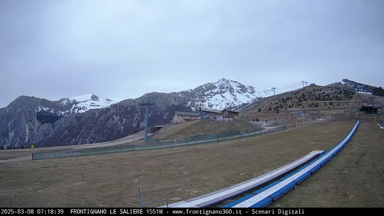 A snow-capped mountain range is seen under an overcast sky, with a ski lift and grassy area in the foreground.