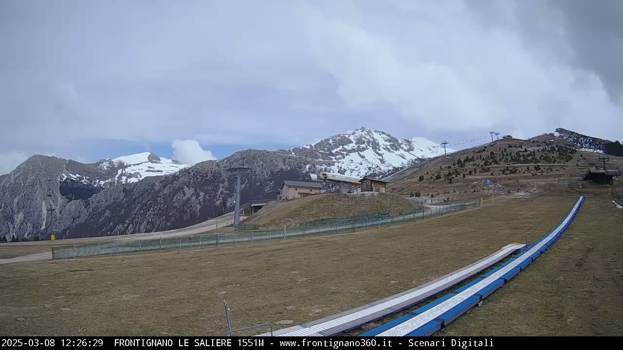 A mostly cloudy day reveals snow-capped mountains and a ski slope with lift infrastructure in a mountainous region.