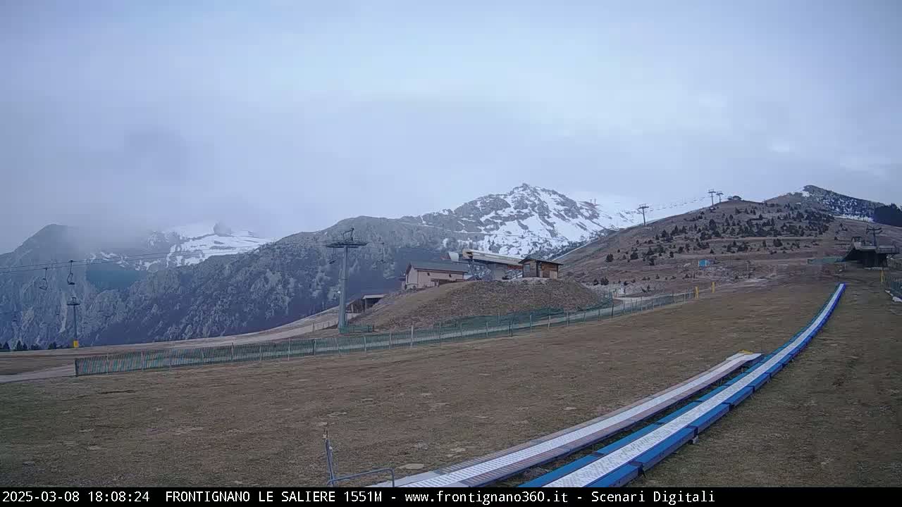 A mostly cloudy, overcast day shows snow-capped mountains in the background, with a ski lift and slope in the foreground.