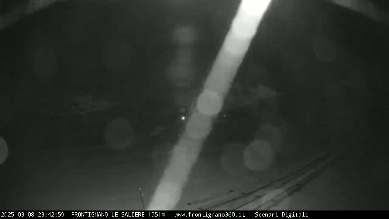 A nighttime, blurry, low-light image shows a road or path with some indistinct objects and a bright light streak across the scene, possibly from rain or snow.