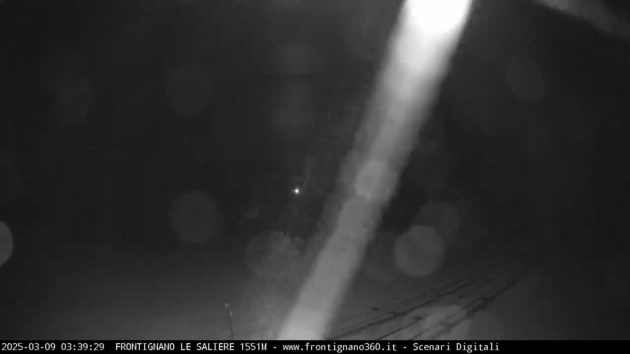 A blurry, nighttime image shows a bright light source streaking across a dark scene with indistinct objects and faint lights in the background.
