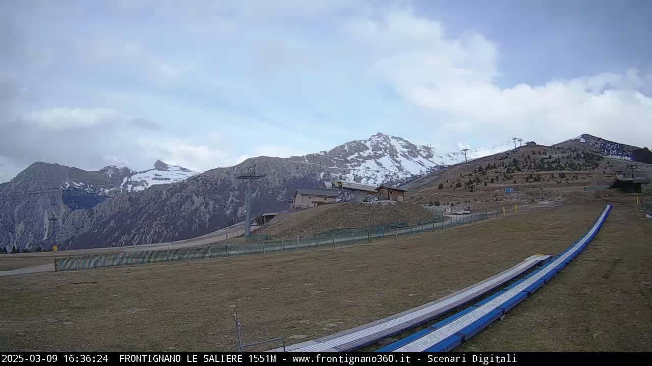 A mostly cloudy day reveals snow-capped mountains in the background, with a ski lift and slope in the foreground.