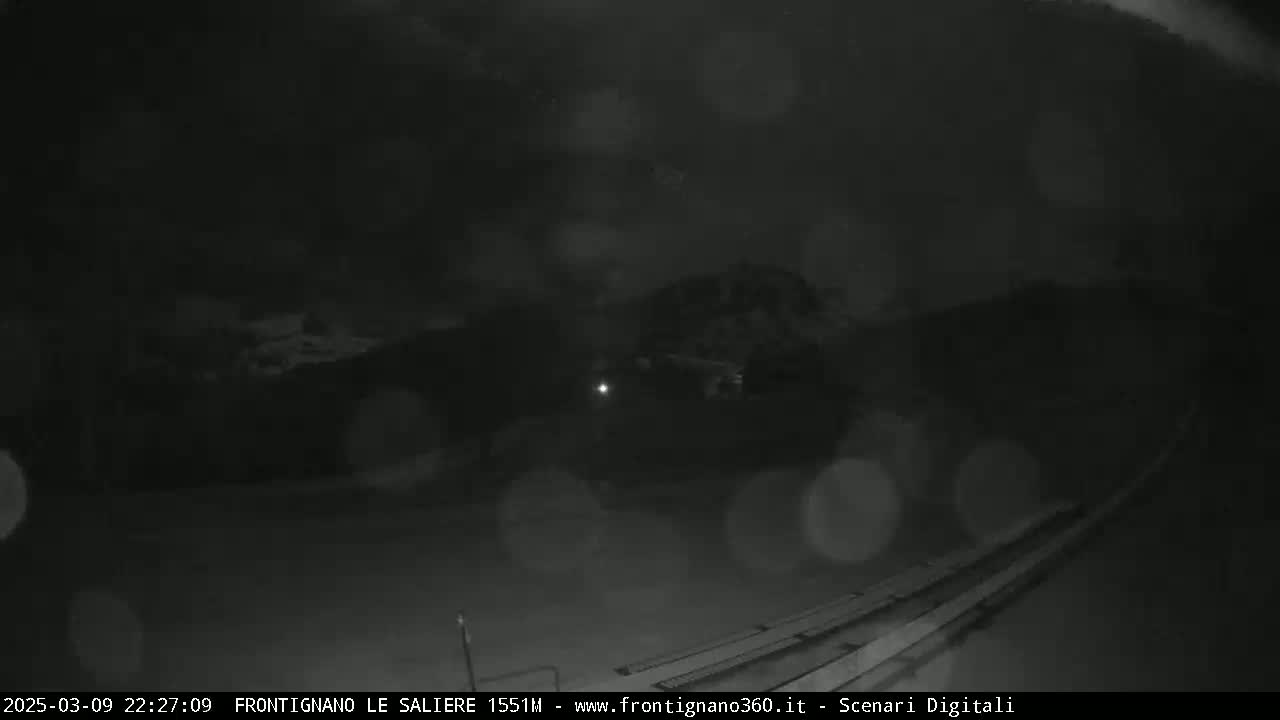 A nighttime, blurry, low-light image shows a dark landscape with a few lights and what appears to be rain or snow.