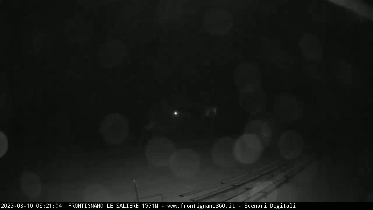 A nighttime, snowy scene shows a dimly lit road with tracks and blurry lights in the distance.