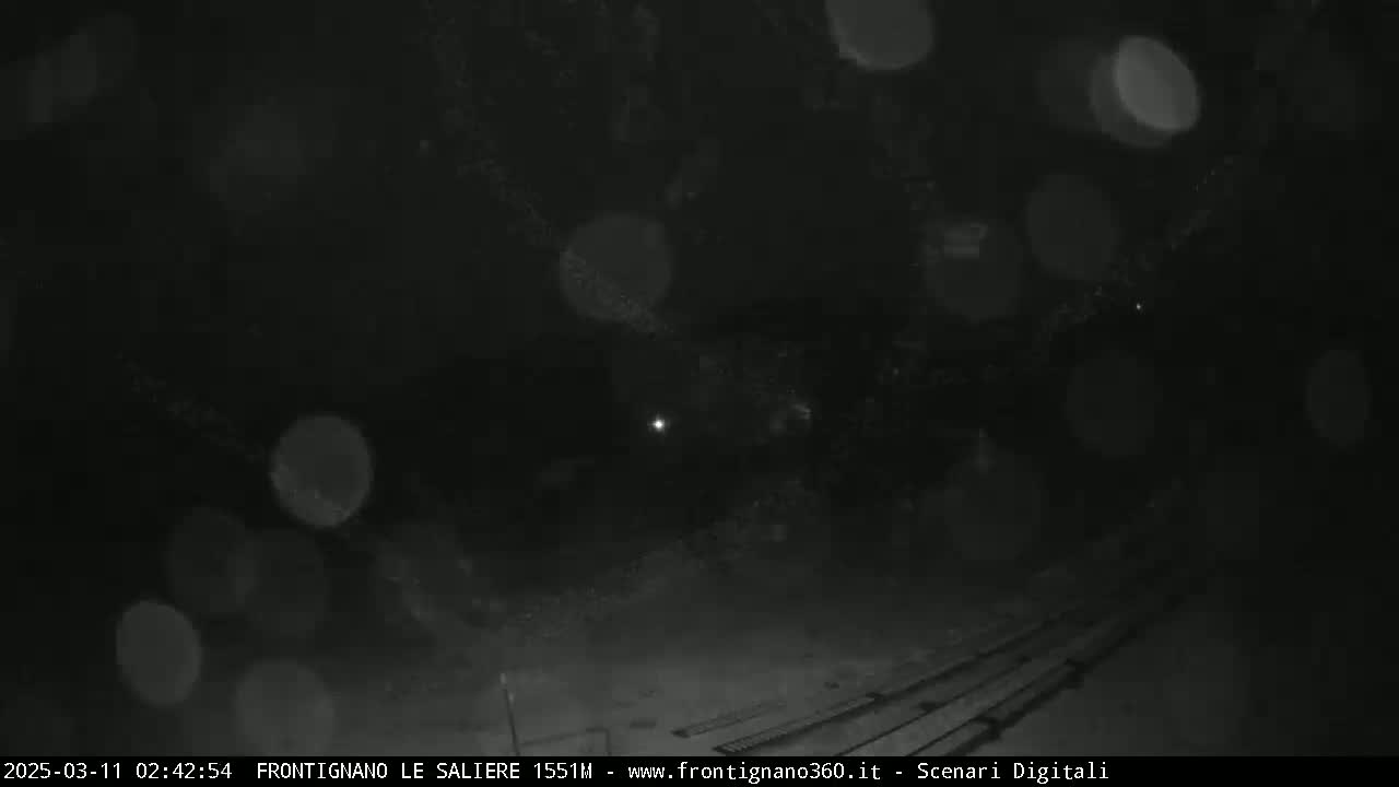 A nighttime outdoor scene shows a dark area with some faint lights and blurry, bright spots possibly from falling snow or rain.