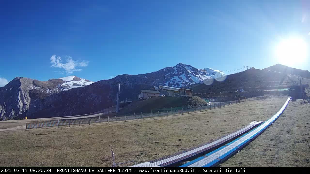 A sunny day in a mountainous region shows snow-capped peaks, ski lift infrastructure, and a long, empty ski lift conveyor belt in the foreground.