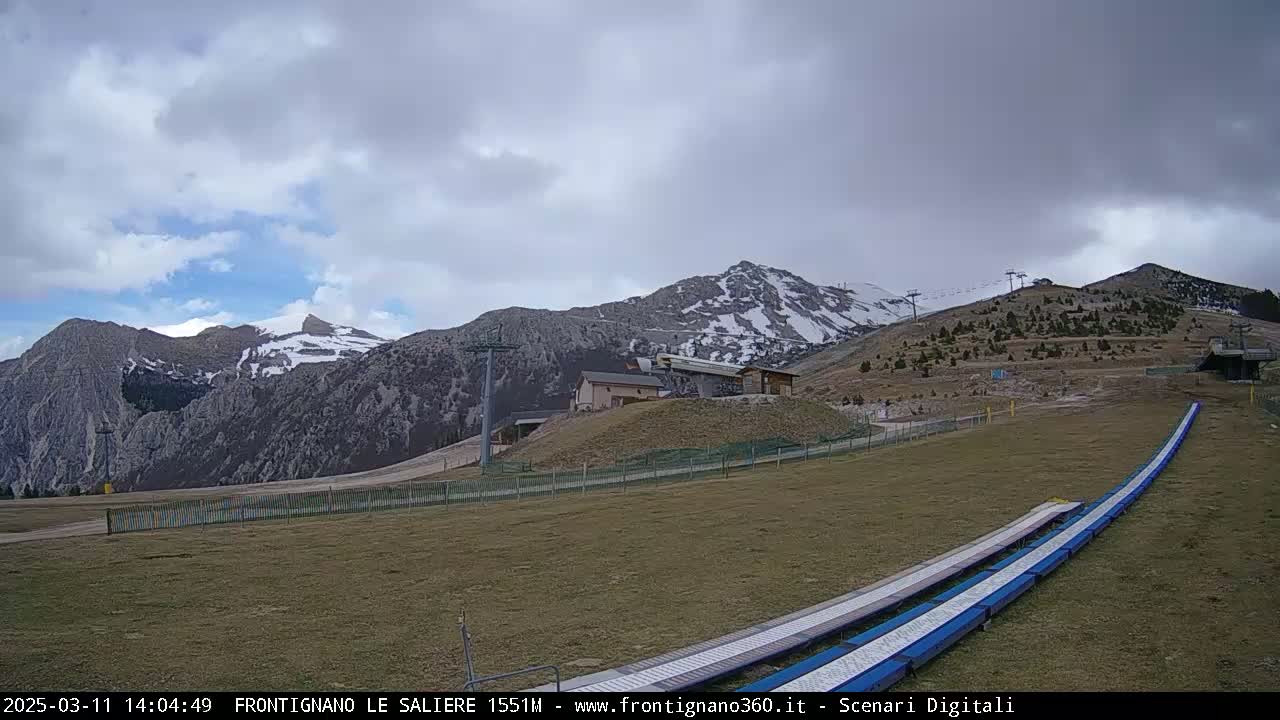 A mostly cloudy day reveals snow-capped mountains in the distance, with ski lifts and grassy slopes in the foreground featuring two long, blue ramps.
