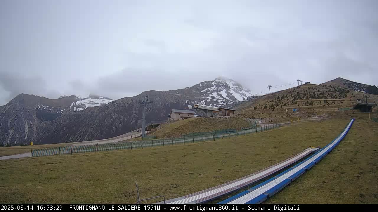A mostly cloudy day reveals snow-capped mountains in the background of a grassy area with ski lifts, a small building, and two long, flat structures possibly used for recreational activities.
