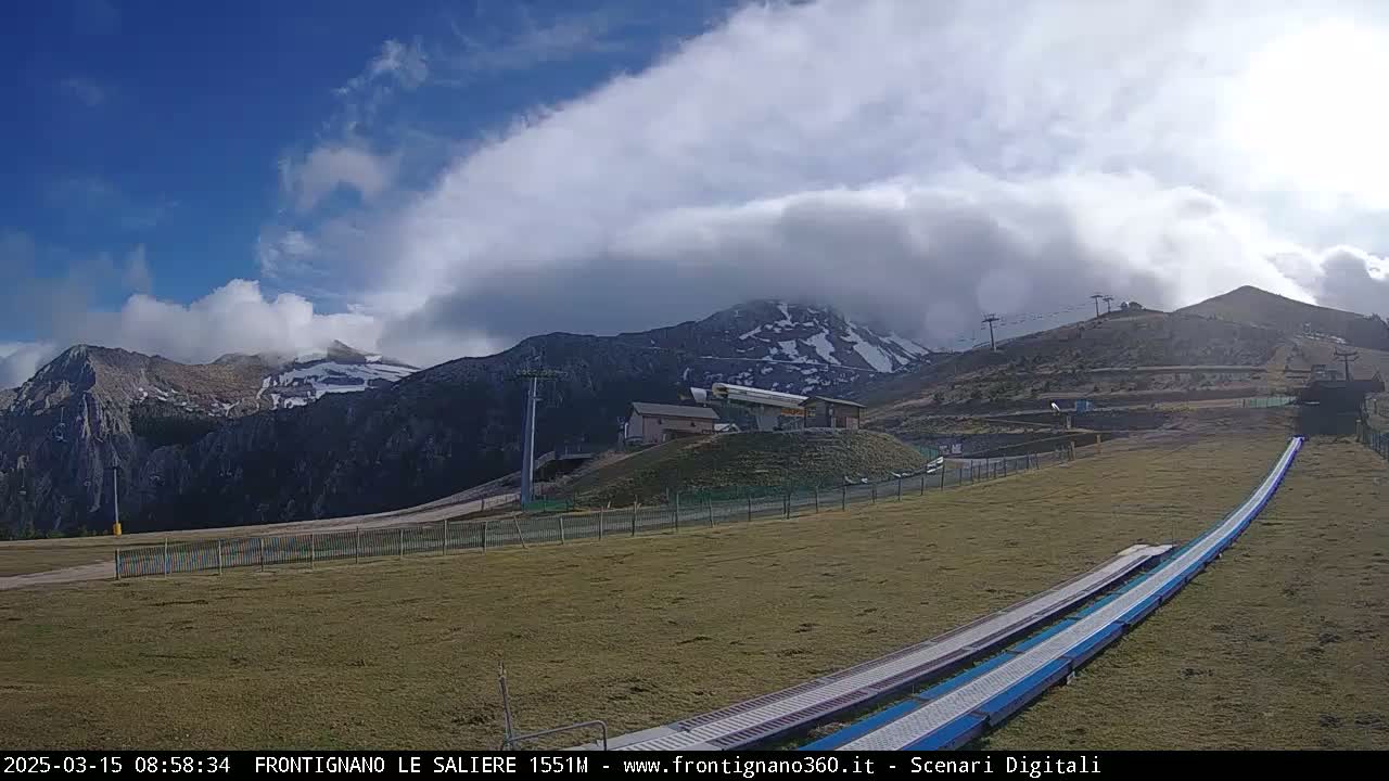 A mountain ski resort with a chairlift and grassy areas under a partly cloudy sky.